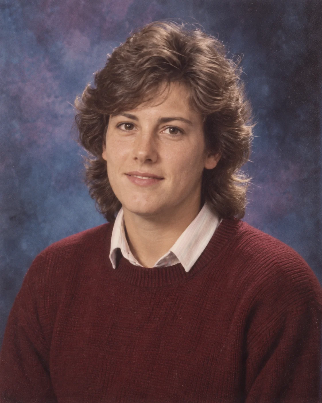 Person with feathered brown hair wearing a burgundy sweater against a mottled blue yearbook backdrop