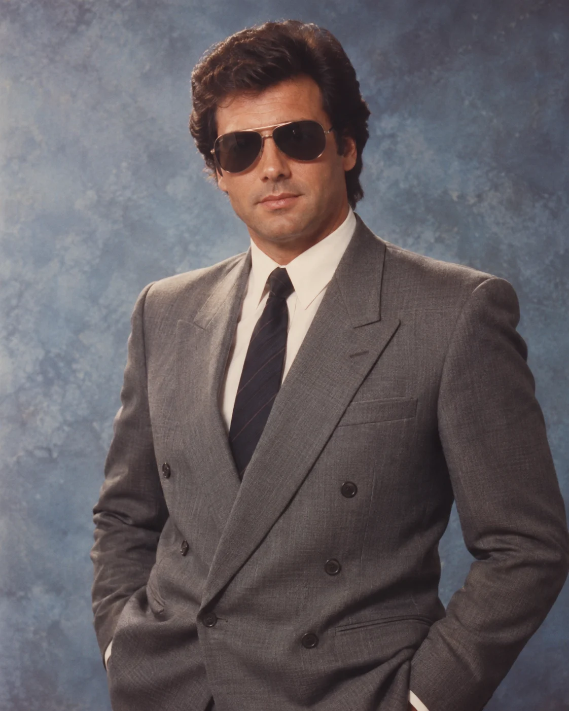 Man in a gray double-breasted suit and aviator sunglasses against a mottled blue studio backdrop